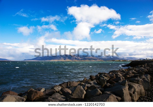 \
View of the Atlantic Ocean and the mountain\
covered with clouds from the embankment of Reykjavik on a sunny but\
windy day. Large stones divide the ocean and the embankment of the\
capital of Iceland