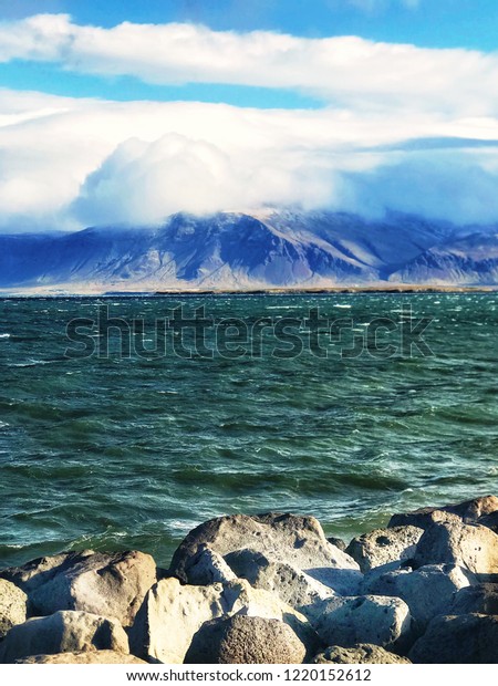 View of the Atlantic Ocean and the mountain\
covered with clouds from the embankment of Reykjavik on a sunny but\
windy day. Large stones divide the ocean and the embankment of the\
capital of Iceland