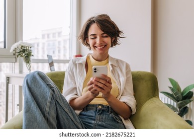 View of attractive woman using her phone .Brunette carre caucasian woman smiling and looking at phone sitting on the chair in room. Concept use technology