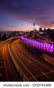 View Of Auckland City Skyline, Sky Tower, And Motorway With Car Trails At Sunrise.