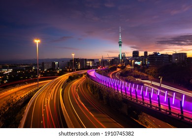 View Of Auckland City Skyline, Skytower, And Motorway With Car Trails At Sunrise.