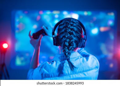 View from back of beautiful gamer woman playing video game at home in front of big screen with joystick and headphone. Colorful neon led. - Shutterstock ID 1838245993