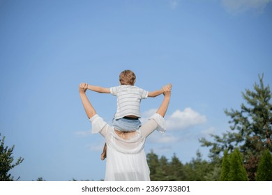 View from back, boy is sitting on his mother's shoulders, their hands are joined against blue sky. Single parents. Family relations. Happy together. Love and understanding. Holidays invillage.