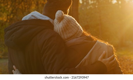 View from back close-up young couple in love in autumn park hugging cute chatting talking enjoying pleasant pastime together admiring view of sunset basking in last warm sunbeams standing in embrace
