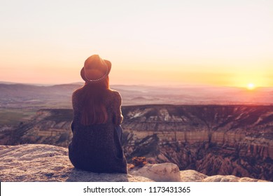 A view from the back of a girl in a hat sits on a mountain and admires the beautiful view and dawn or sunset in Cappadocia in Turkey. - Shutterstock ID 1173431365