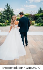 View From The Back. The Groom In A Classic Suit And The Blonde Bride In A White Dress Walk Together Holding Hands. The Concept Of Love, Mutual Understanding And Happy Family Life.