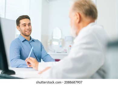 View From Back To Happy Young Man Patient Listening Senior Male Doctor Giving Healthcare Advice During Checkup Visit In Clinic Office. Senior Physician Interviewing Smiling Young Man At Hospital.