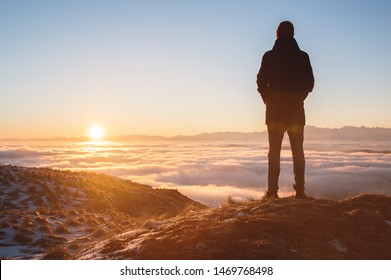 View From the back. A lonely standing man high in the mountains looks at the setting sun and the sunset horizon with a valley filled with clouds. The concept of tourism travel and male loneliness - Powered by Shutterstock