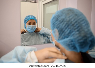 View From The Back Of A Medical Professional, Doctor, Nurse, Surgeon In Protective Workwear And Medical Mask Looking At His Mirror Reflection In Dressing Room Of Medical Surgery Clinic