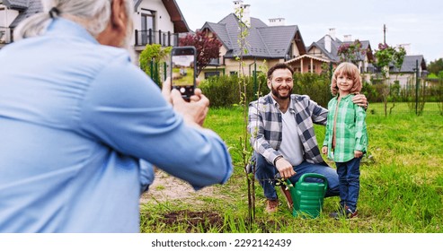 View from back on grandfather taking photo with smartphone of happy father and cute small son smiling and giving thumbs up after planing trees in garden. making picture with phone. Family at house. - Powered by Shutterstock