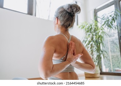 View From Back On Senior Female In Top Stretching Arms, Keeping Hands Behind Back, Doing Yoga Exercises. Healthy Lady Sits On Fitness Mat At Home