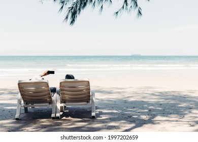 View From The Back Of A Retired Couple Resting On A Adirondack Chair By The Beach In The Sunlight And Clean Sandy Beach. Retirement Planning Ideas And Happy Life.