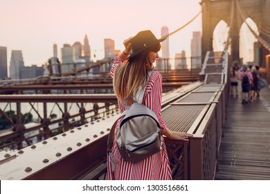 View From Back Of  Traveling Woman In Red Dress, Stylish Black  Hat And Silver Bag Pack Enjoying Amazing View From Brooklyn Bridge In New York. Sunset Colors.