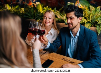 View from back of unrecognizable female clinking glass of red wine with smiling beautiful man and woman sitting at table in fancy restaurant at evening. Happy three friends enjoying nice dinner.
