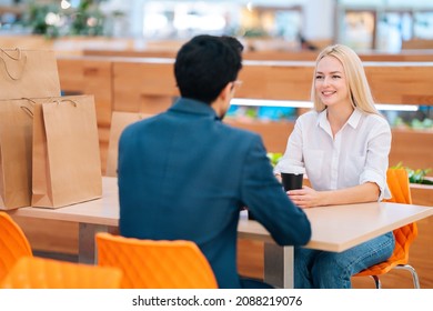 View from back to unrecognizable man and attractive blonde woman having date sitting at table looking at each other in mall. Beautiful young couple drinking coffee and talking in cafe after shopping.