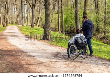 View from back to a woman in wheelchair and a man walking and checking his phone in a park near river at spring. Woman with a disability in wheelchair and her personal assistant are hiking together