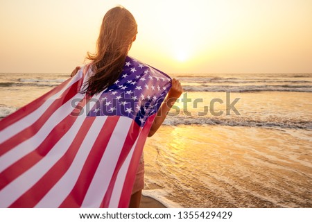 view from the back young brunette female person holding national American flag cover ocean beach holiday travel at summer romantic sunset .America independence day concept
