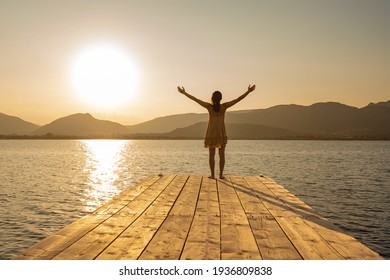 View from back of young woman on wooden pier in short dress and barefoot with open arms in contemplation of the sun setting among the mountains on lake or ocean sea. Orange vintage effect photography - Powered by Shutterstock