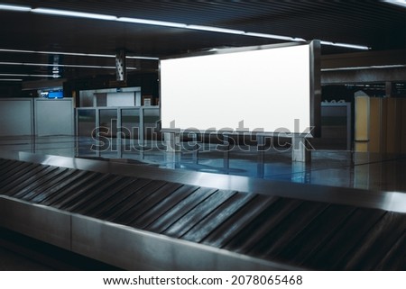 View of a baggage claim area in a hall of a modern airport arrival zone with luggage conveyor belt, with a selective focus on a template of a blank advertising poster or an information billboard