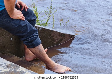 View Of Bare Male Feet In The Water. A Man Cools His Feet On The Steps Of The Descent Of The Embankment Of The River During The Flood. Jeans Are Rolled. The Concept Of Relaxation By The Water.