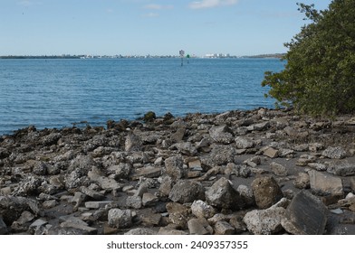 View  of bay on bayside at Jungle Prada de Narvaez Park looking west in St. Petersburg, Florida on a sunny day. Rocks on the shoreline. Green bushes on right. Blue sky with white clouds and calm water
