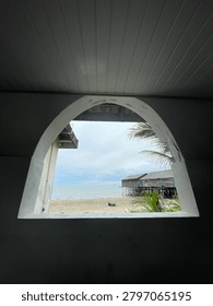 View of a beach through an arch-shaped window with palm trees and shoreline.