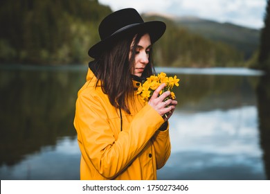 View of a beautiful girl with hat and yellow jacket looking at view and smelling wild flowers. Thoughtful stylish female enjoys the outdors with mountains and lake on background. Travel and wanderlust - Powered by Shutterstock