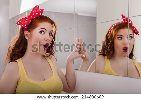 View of a beautiful redhead girl with colorful pinup clothes next to a mirror wall.