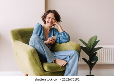 View of beautiful woman in home with phone .Brunette carre hair look and smiling to camera sitting on the green chair at home. Relax, lifestyle concept  - Shutterstock ID 2182675191