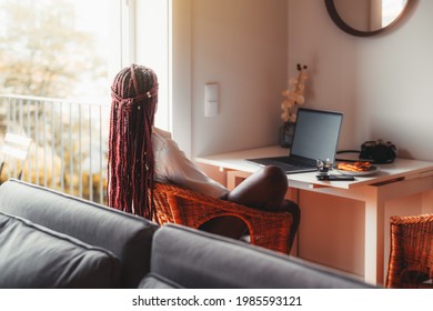 View From Behind Of A Black Woman With Long Hanging Braids Sitting In The Living Room Of Her House In A Cane-chair And Pensively Looking Outside The Window, A Laptop And Morning Coffee On The Table