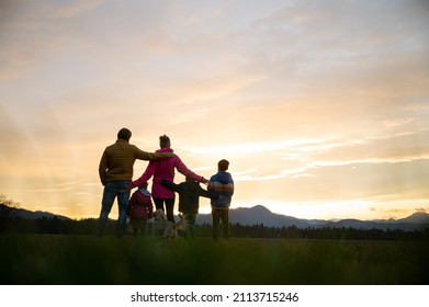 View From Behind Of Family Of Five And A Dog Standing Outside At Dusk With Teir Arms Around Each Other.