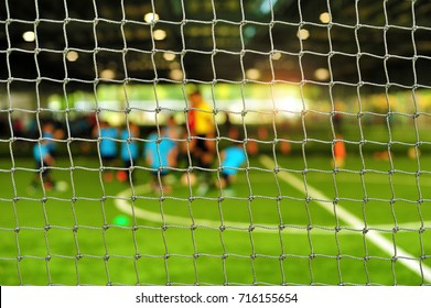View From Behind The Goal Of Soccer Field. Blurry Scene Of Football Coach And Children Playing Soccer In Football Field At Indoor Stadium. Soccer Training For Kids.