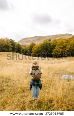 View from behind. A hiker with a backpack and hat walks along a rocky mountain trail. The young woman enjoys the mountain scenery and feels a sense of freedom. Concept of hiking, nature, and travel.
