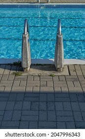 A View From Behind A Ladder At The Edge Of An Outdoor Public Pool Shows Its Railings Supported By Concrete.