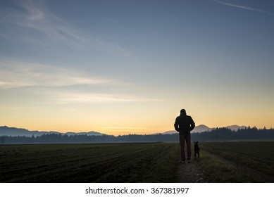 View From Behind Of A Man Walking With His Black Dog At Dusk On A Country Road Leading Through Beautiful Landscape Of Fields With Forest In The Distance And Beautiful Evening Sky Above.