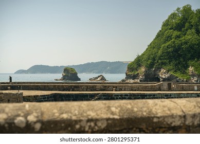 A view from behind a stone wall overlooking the harbour wall with a lone figure fishing towards small sea stacks and a densely wooded cliff face in Charlestown, Cornwall, United Kingdom.
