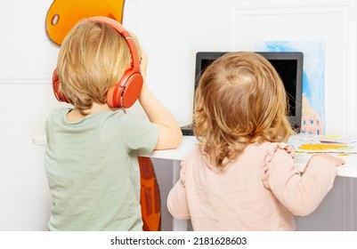 View From Behind Of Two Kids, Boy And A Girl Watch Education Videos Over Internet On Laptop Wearing Headphones