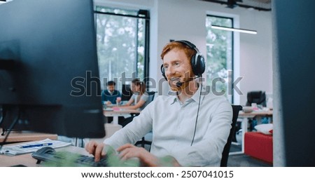 View from behind of two large monitors of Caucasian male sitting at desk Smiling while talking with customer. Man working hard in big office. Wear on wired headset with attached microphone.