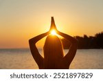 View from behind of a woman with long hair standing by the sea practicing mindfulness and gratitude. Holding her palms joined above her head with sunlight of the rising sun.