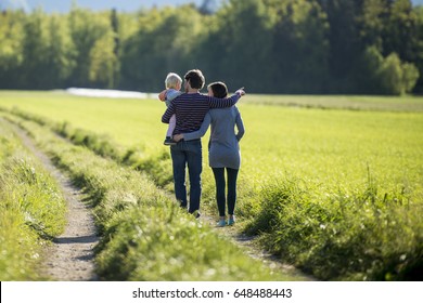 View From Behind Of A Young Family On A Country Road Surrounded By Green Meadow.