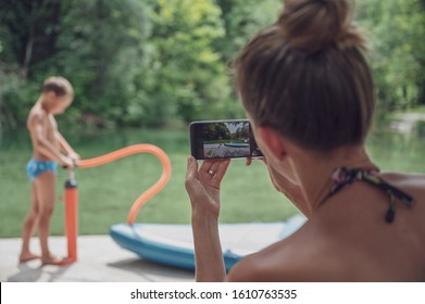 View From Behind Of A Young Mother Taking A Photo Of Her Son By The River With Her Mobile Phone.