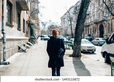 View From Behind Young Woman In Black Coat And With Curly Hair Walking Through An City Street Next To Road And Cars On Autumn Day.