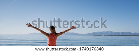 View from behind of a young woman in pink dresds standing by the calm morning sea with her arms spread widely as she greets the new day. Wide view image.
