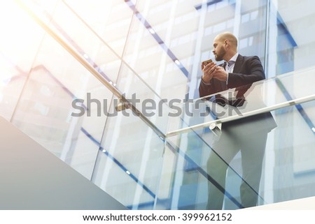 View from below of a businessman is holding mobile phone in hands, while is standing in skyscraper interior with modern design background with copy space for your advertising text message or content