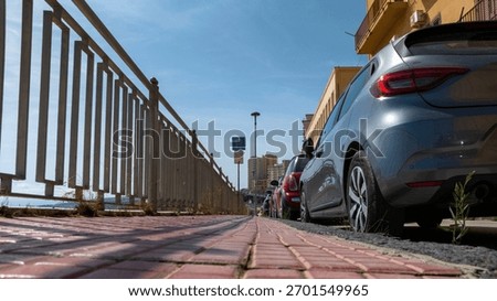 A view from below of an empty sidewalk. On one side is a metal railing, on the other side are parked cars.