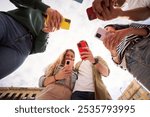 View from below of group young people standing in circle holding colorful mobiles phones outdoors. Generation z friends addicted to technology and social networks using cells together on sunny day 