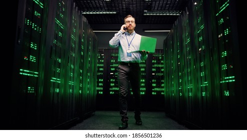 View from below on Caucasian man walking among servers with laptop computer and talking on mobile phone. Male analytic in data storage working on digital security and speaking on cellphone.