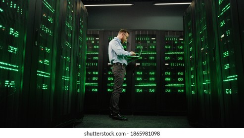 View from below on Caucasian man walking among servers with laptop computer and checking big data processors. Male analytic in data storage working on digital security and safe telecommunication.