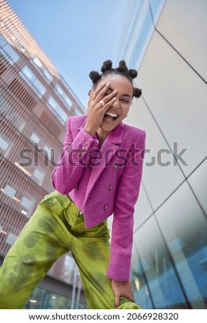 View from below of overjoyed female model keeps hand on face feels very happy wears stylish pink jacket and green trouses spends free time in urban place expresses positive emotions. Youth style