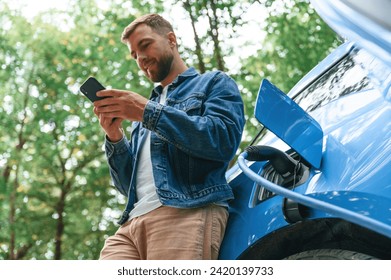 View from below, with smartphone. Handsome man is waiting for his electric car to charge, outdoors. - Powered by Shutterstock